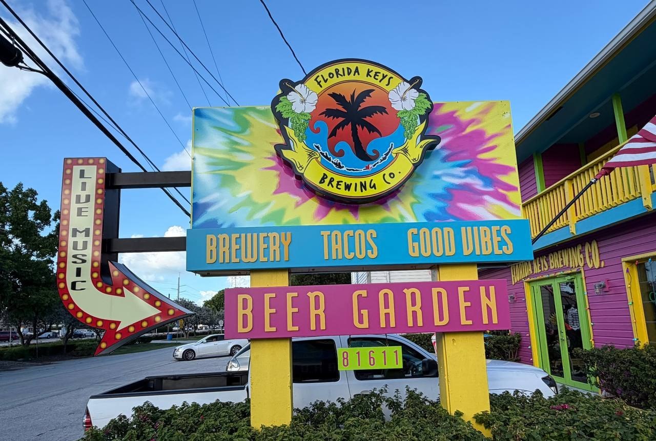 Tie-dye Florida Keys Brewing Co. sign with Brewery, Tacos, Good Vibes and Beer Garden panels beside a lit Live Music arrow