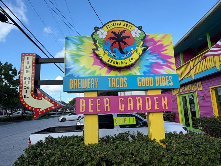 Tie-dye Florida Keys Brewing Co. sign with Brewery, Tacos, Good Vibes and Beer Garden panels beside a lit Live Music arrow