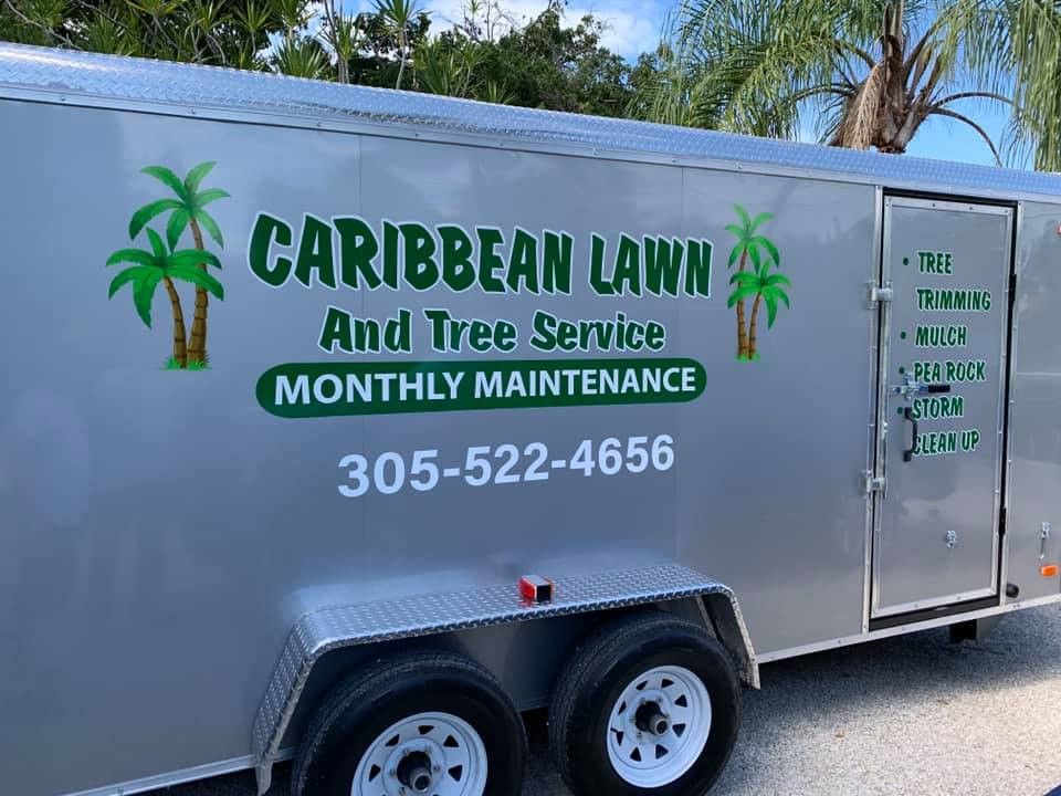 Silver enclosed trailer lettered Caribbean Lawn and Tree Service with palm trees and a phone number